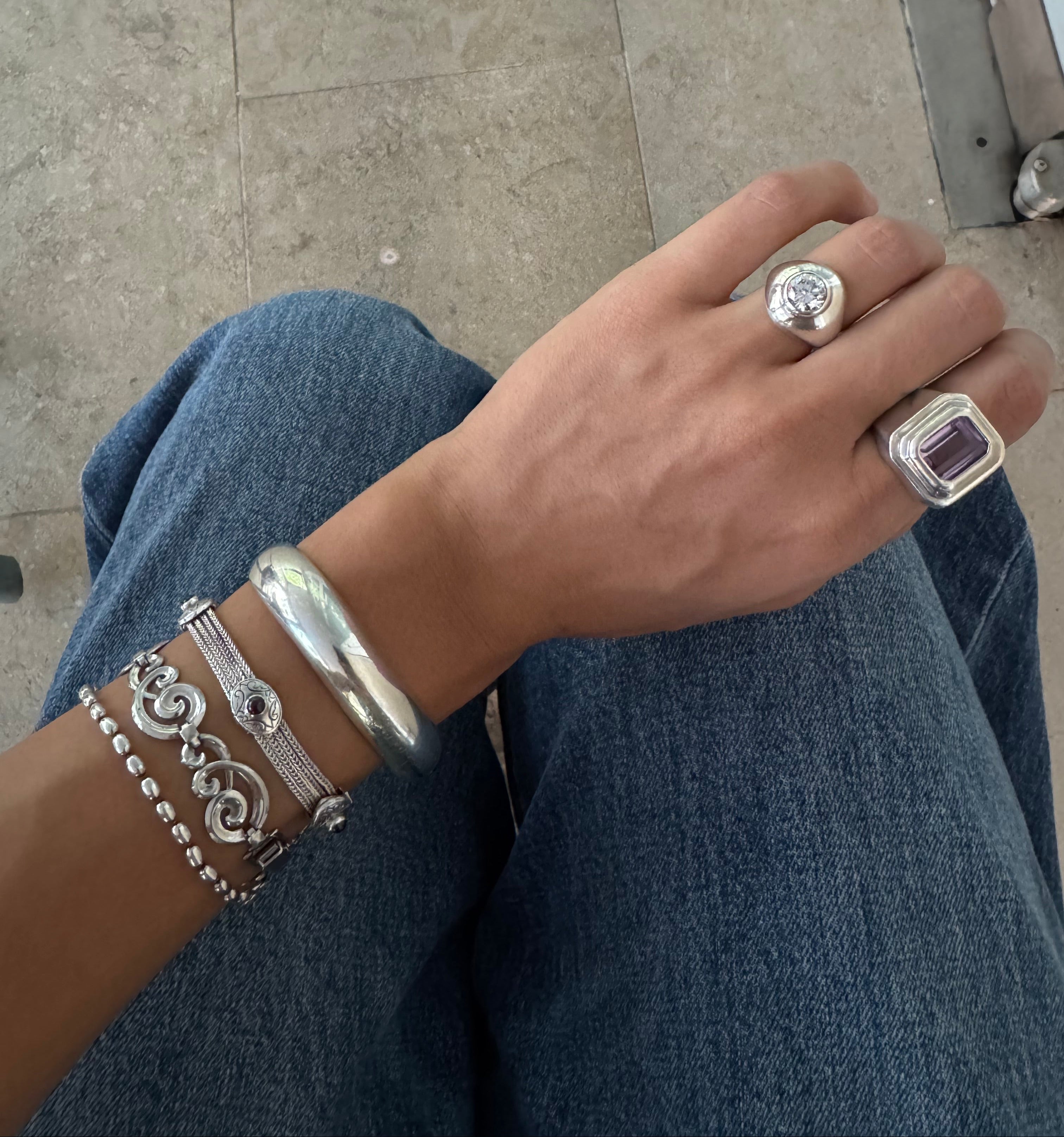 A close-up of a person's hand wearing multiple silver-toned jewelry pieces, including a large signet ring with a purple rectangular stone, a smaller silver ring, and stacked silver bracelets, while sitting on blue jeans with a tiled floor visible in the background.