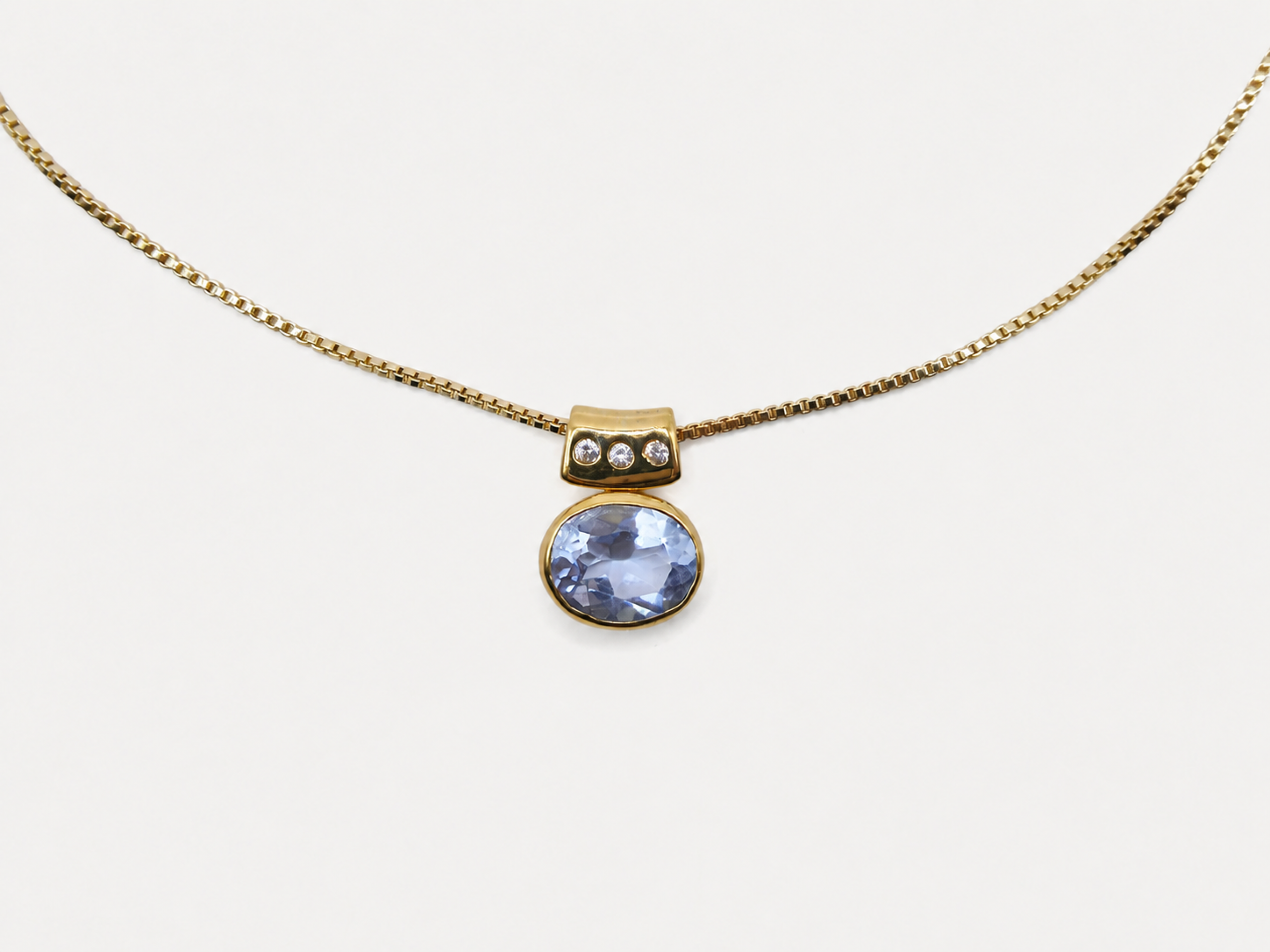 A gold chain necklace with a pendant featuring a round light blue gemstone and a small gold cap with three tiny clear stones, shown on a plain light background.