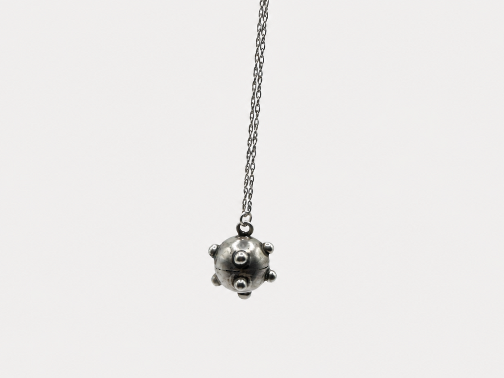 A silver-toned pendant necklace featuring a small, spherical, spiked ball charm hanging from a delicate chain, set against a solid light gray background.