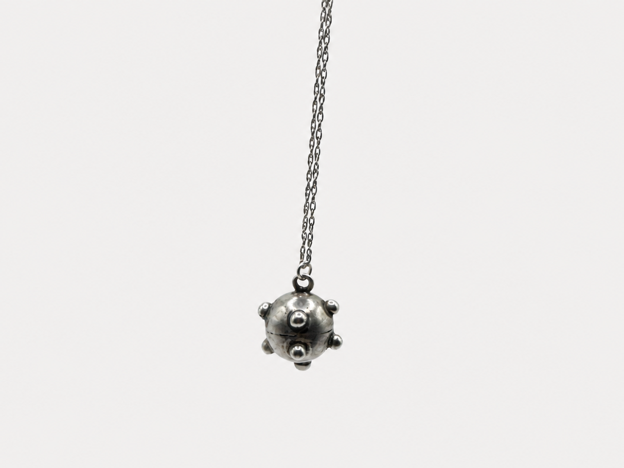 A silver-toned pendant necklace featuring a small, spherical, spiked ball charm hanging from a delicate chain, set against a solid light gray background.